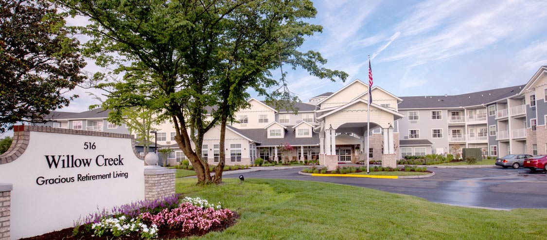 A large, three-story white building with an American flag at the entrance, surrounded by manicured lawns and colorful flower beds. A sign in the foreground reads 