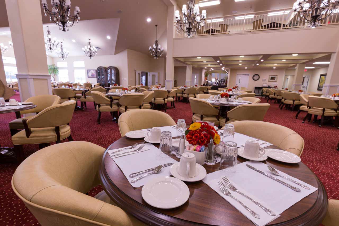 A spacious dining room with elegant chandeliers, round tables set with white dishes, glasses, and silverware. Each table features a centerpiece of vibrant red and yellow flowers. Beige upholstered chairs surround the tables, and the room is carpeted in red.