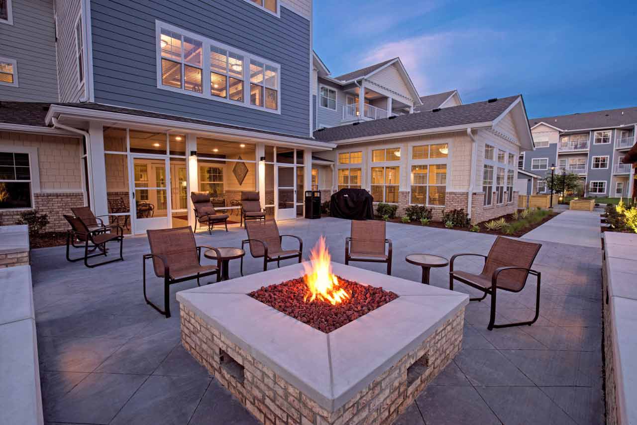 A modern outdoor patio featuring a stone fire pit with a bright flame, surrounded by six brown chairs. The patio is part of a larger building with windows and includes neatly landscaped pathways and greenery. The evening sky adds a serene atmosphere.