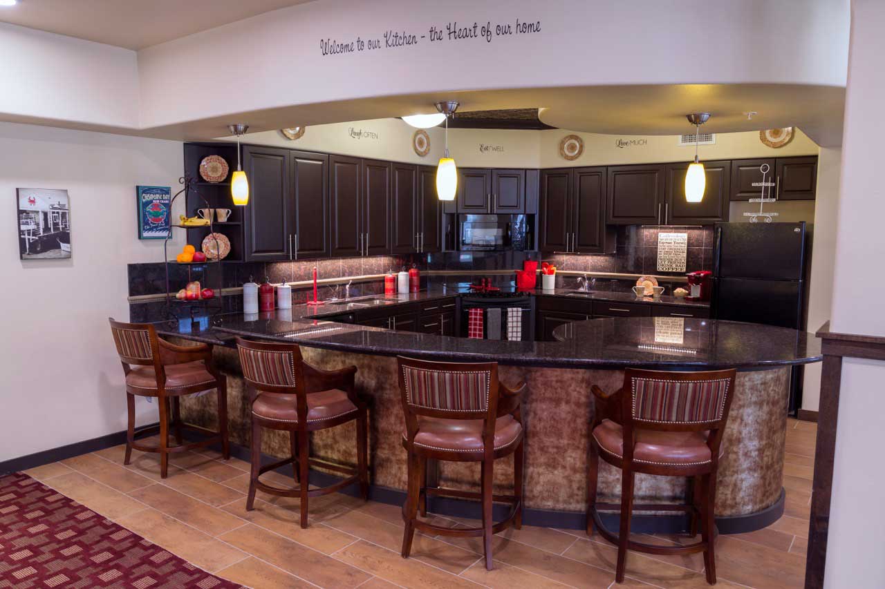 A modern kitchen with dark wood cabinets, a large granite countertop island with four wooden chairs, and various nautical-themed decorations. The wall above the kitchen reads, 