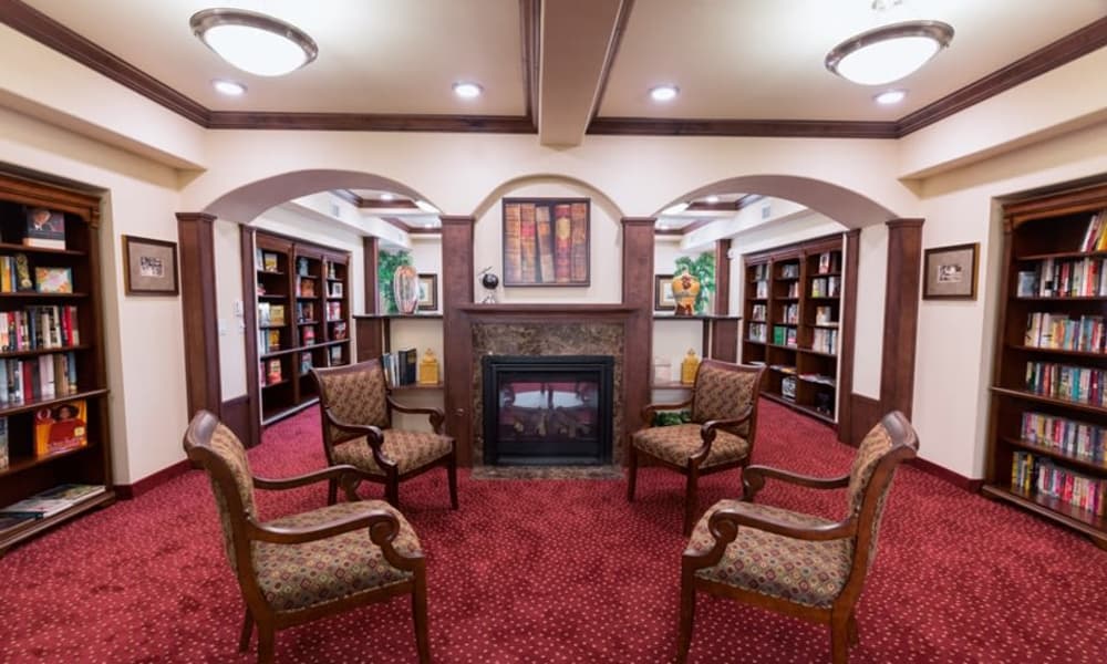 A cozy, well-lit library with red carpeting and wooden bookshelves lining the walls. Four upholstered wooden chairs are arranged in front of a fireplace. Paintings and plants decorate the space, creating a warm and inviting atmosphere for reading.