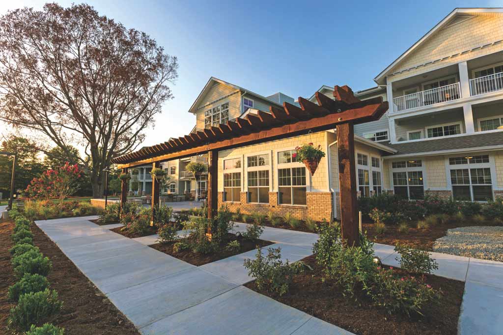 A modern building with a spacious, well-maintained front yard featuring a wooden pergola adorned with hanging plants. The area is landscaped with flower beds and shrubs, and the sun casts a warm glow on the facade, highlighting large windows and balconies.