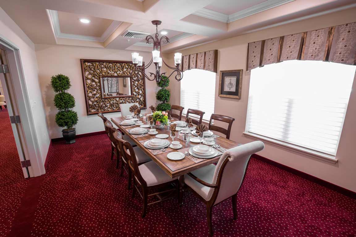 A well-decorated dining room with a wooden table set for eight people, featuring plates, glasses, and a centerpiece. The room has red carpeting, windows with patterned curtains, a chandelier, potted plants, and a large framed mirror on the wall.