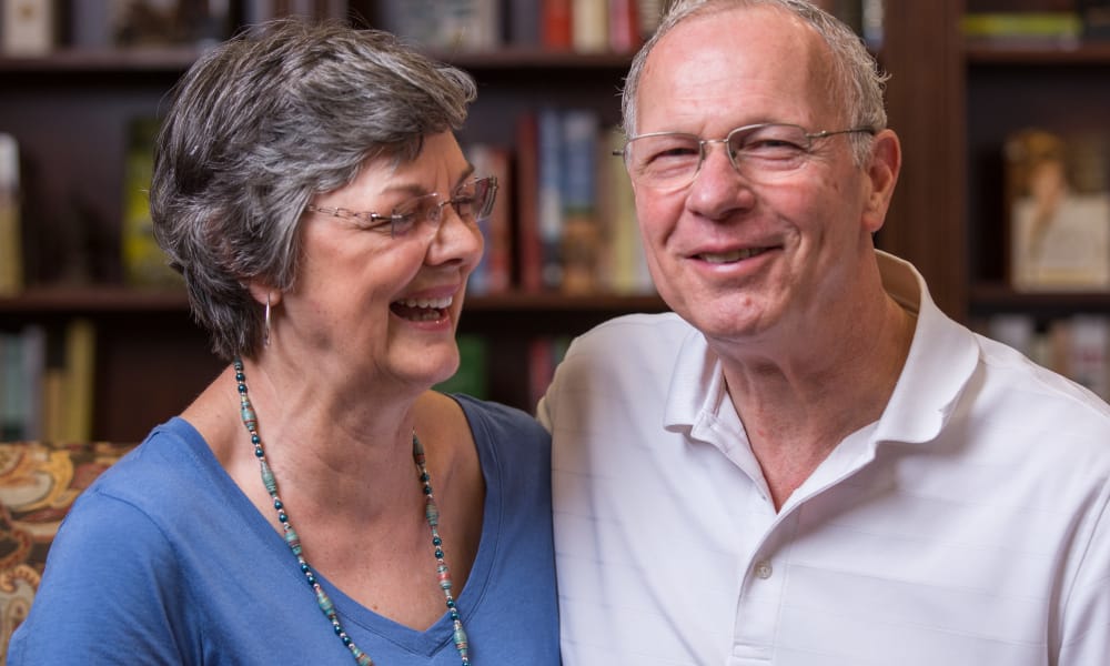 A smiling older couple, with the woman wearing glasses, a blue top, and a beaded necklace, and the man wearing a white polo shirt, sit close together in a cozy room with bookshelves in the background.