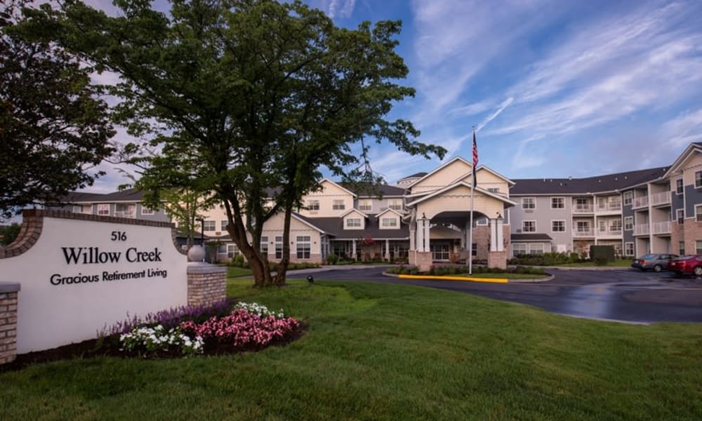 An exterior view of a retirement residence named Willow Creek, featuring a welcoming main entrance with a covered driveway. The building has multiple stories, landscaped grounds, and an American flag. The sign in front reads 