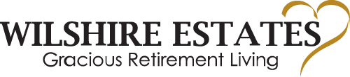 Logo for Wilshire Estates with the tagline "Gracious Retirement Living." The text is in a serif font with the "i" in "Wilshire" dotted by a pair of interconnected heart shapes in gold.