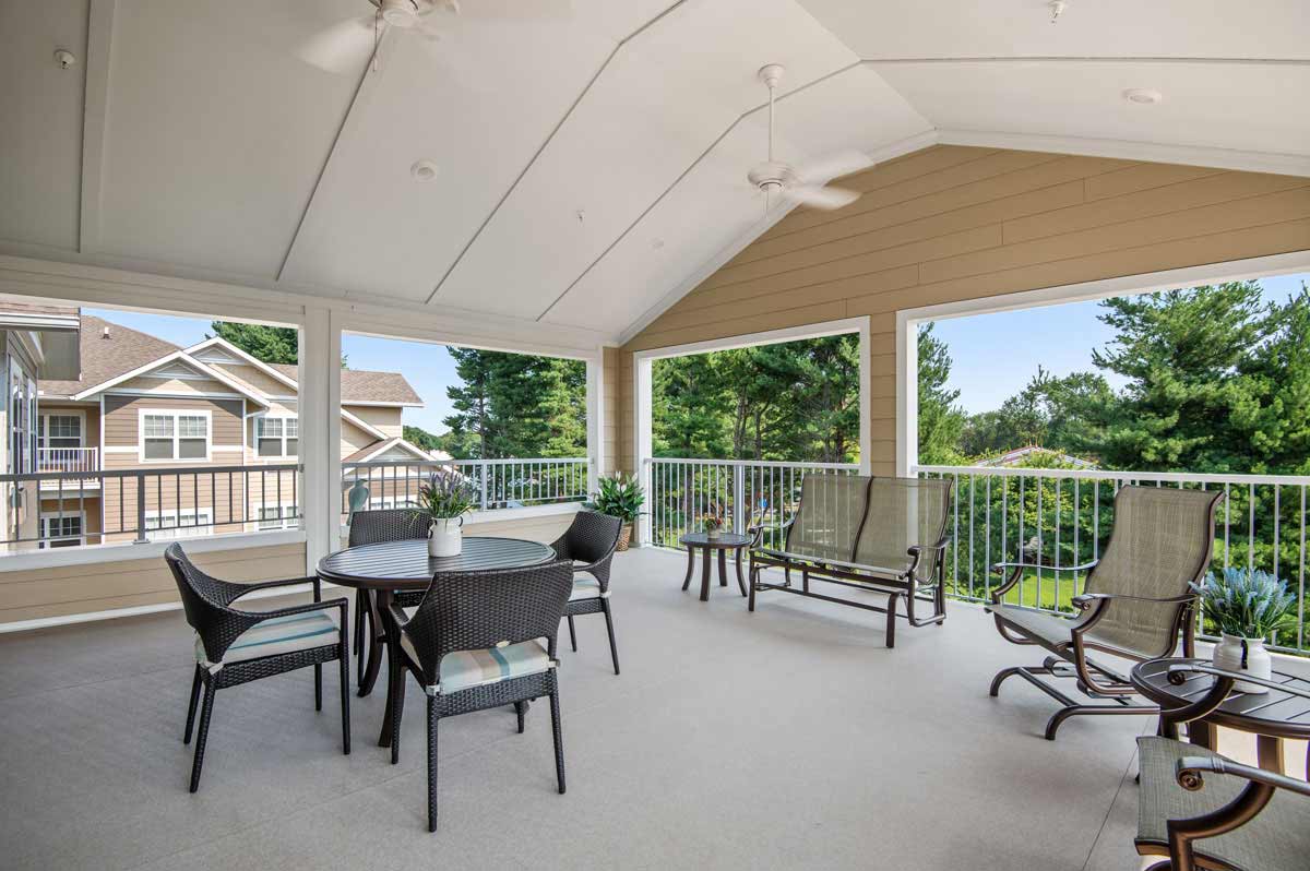 A spacious covered patio features a round table with four wicker chairs, two lounge chairs, and two rocking chairs. Potted plants adorn the corners. The area overlooks a well-maintained garden and neighboring buildings under a clear sky.