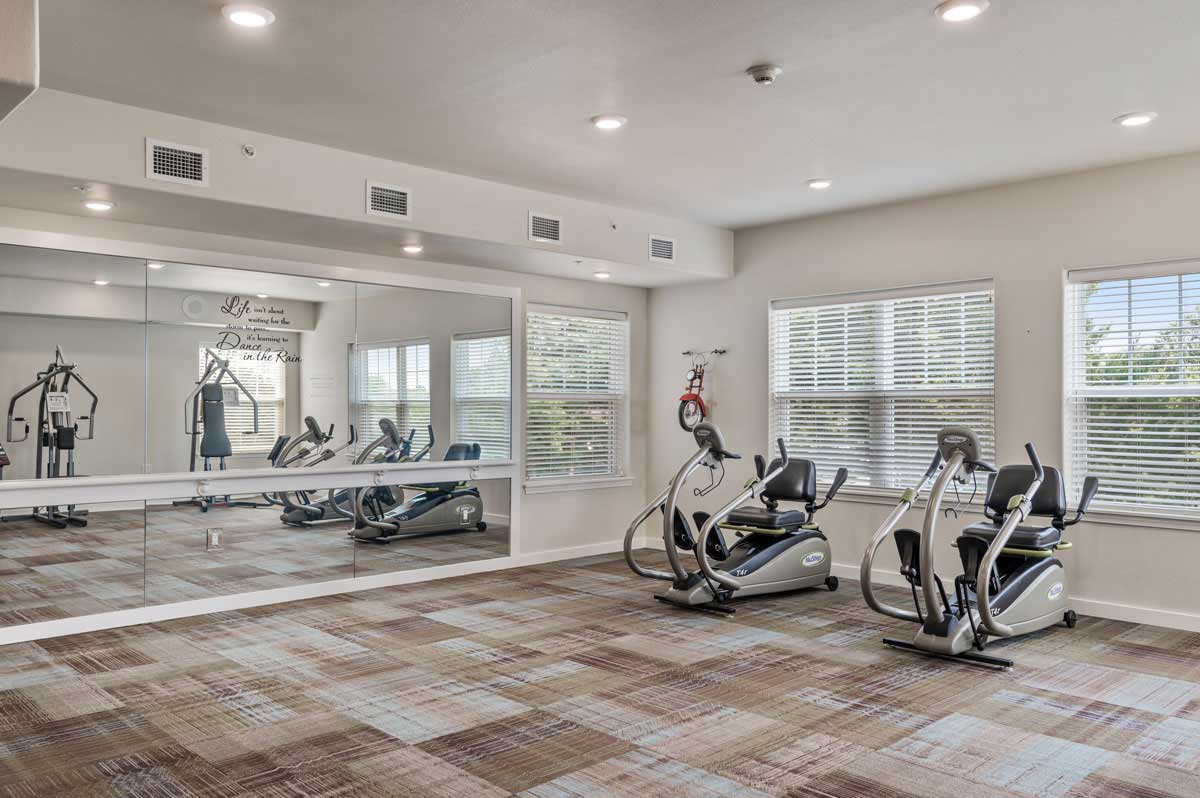 A modern fitness room with large mirrors, carpeting, and natural light from windows. Features include two recumbent exercise bikes and a weight machine. The room has overhead lighting and motivational quotes on the wall.