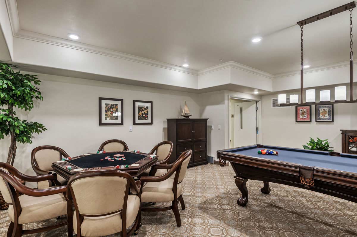 A spacious game room featuring a poker table with five chairs on the left and a pool table on the right. The room is well-lit with recessed lighting and a hanging light fixture over the pool table, and has framed artwork on the walls and a green potted plant in the corner.