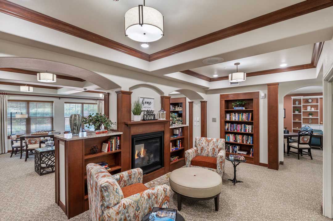 A cozy living room with patterned armchairs around a round ottoman. A lit fireplace is set in a wooden bookshelf unit filled with books and decor items. The space is warmly lit by ceiling lights, and a dining area is visible in the background.