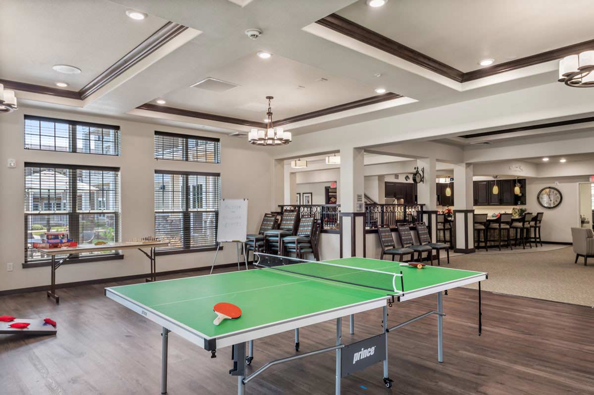 A spacious recreation room features a green ping pong table with a paddle and ball on top, large windows, ceiling lights, and modern decor. In the background, there are tables and chairs, a whiteboard, and a cozy lounge area with a wall-mounted TV.