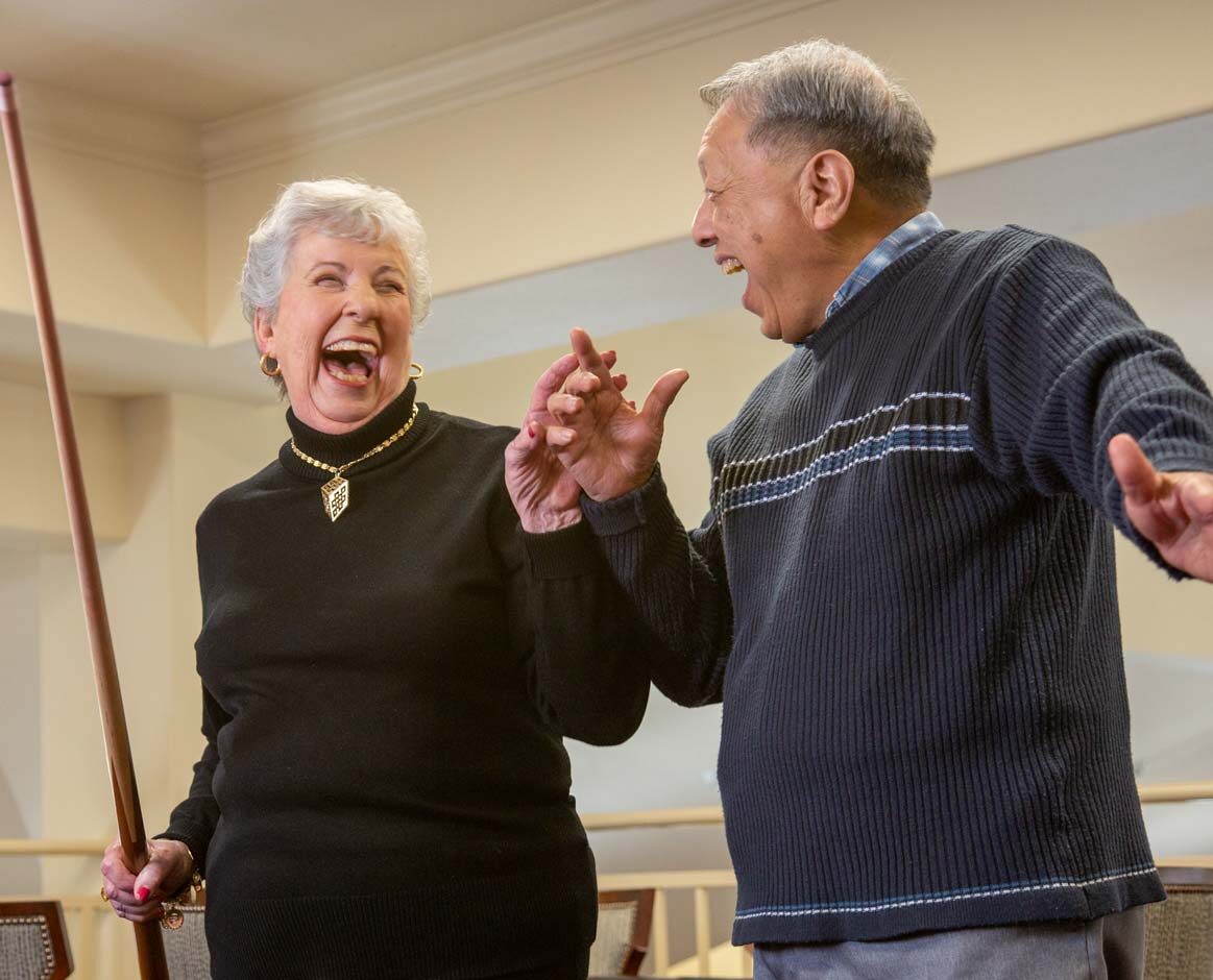 Two elderly individuals are laughing joyfully. The woman, holding a pool cue, and the man, gesturing animatedly, are both engaged in a lively conversation. They are indoors, and the atmosphere is warm and cheerful.