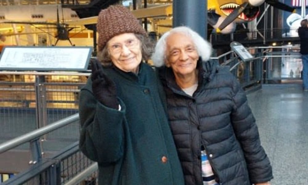 Two elderly women pose together, smiling warmly at the camera. One woman wears a brown knit hat and green coat, while the other wears a black puffer jacket. They stand in an indoor exhibit space with aircraft displays visible in the background.