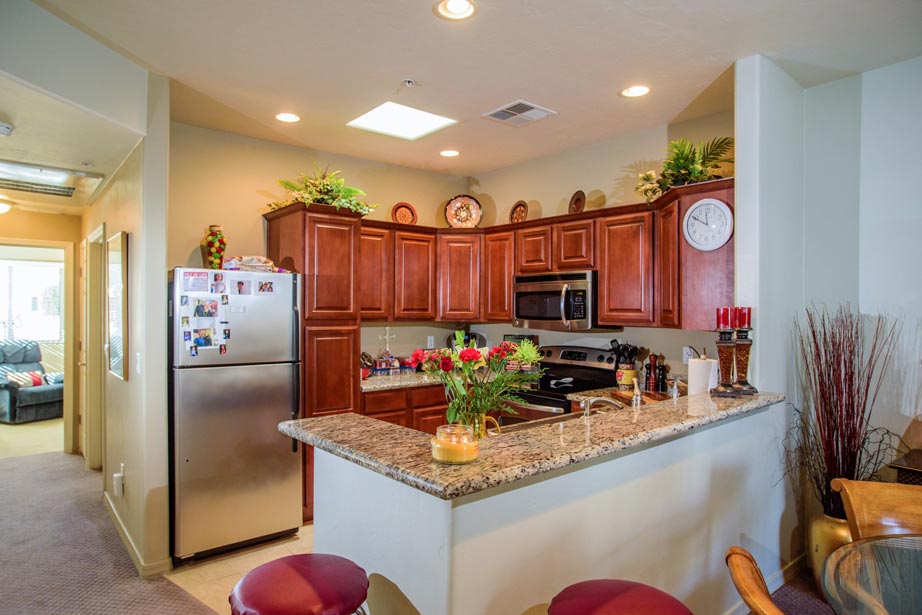A cozy kitchen with wooden cabinets, granite countertops, stainless steel appliances, bar stools, fresh flowers on the counter, and decorative plants and plates above the cabinets. Adjacent living and dining areas are visible.