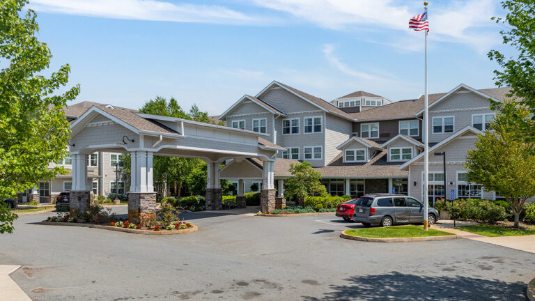 A large, modern building with gray siding and multiple windows, surrounded by greenery. A covered entrance, flagpole with an American flag, and several parked cars are visible in front.