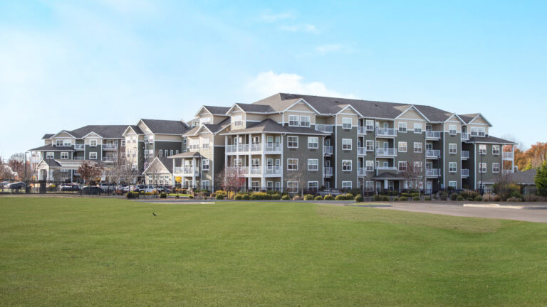 A large, modern apartment complex with green and white exterior sits behind a wide open grassy field under a clear blue sky.