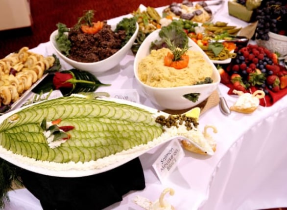 A buffet table with various dishes, including a large platter of sliced cucumbers on a white creamy base, a bowl of seasoned rice, rolls, mixed vegetables, and a tray of fresh fruits like strawberries and grapes, all artfully arranged.