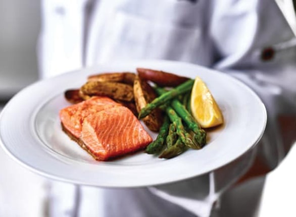 A person in a white chef's uniform holds a plate with a serving of grilled salmon, roasted fingerling potatoes, asparagus, and a lemon wedge. The food is presented in an appetizing and detailed manner.