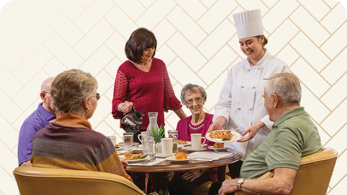 A group of seniors sitting around a table being served by a chef and a woman in a red sweater. The chef is handing over a plate while the woman pours coffee. Plates of food and cups are on the table. The background has a light, tiled pattern.
