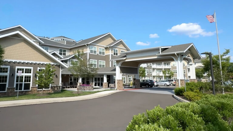 A large, modern assisted living facility with tan siding and white trim, a covered entrance, landscaped bushes, and an American flag flying on a sunny day with blue sky and a few clouds.