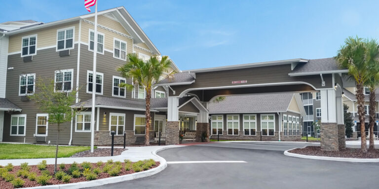 A modern, two-story assisted living facility with gray siding, white trim, and a covered entrance. Palm trees and landscaped greenery surround the building under a clear blue sky.