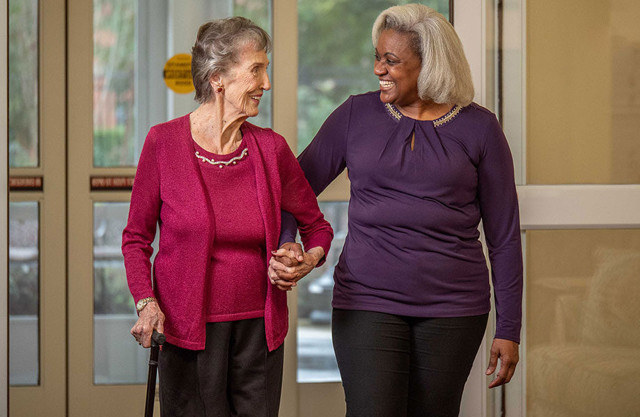 An elderly woman with a cane and a younger woman walk arm in arm, smiling at each other. The elderly woman wears a pink cardigan and black pants, while the younger woman wears a purple top and black pants. They are indoors, near a set of glass doors.