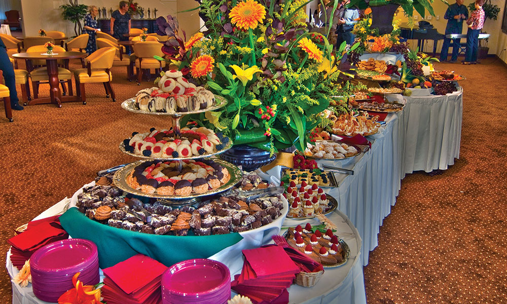 A lavish buffet table is adorned with a variety of desserts and floral arrangements. The table features tiered trays of pastries, cakes, and assorted sweets, surrounded by vibrant flowers and red napkins. In the background, people are seated at tables in a warmly lit room.