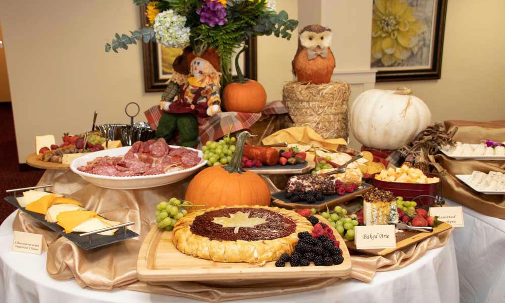 A beautifully arranged autumn-themed banquet table features a variety of cheeses, cured meats, fruits, and pastries. Decorative pumpkins, a hay bale with an owl ornament, and a floral arrangement add festive touches to the display.