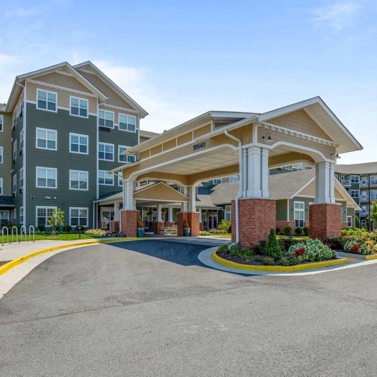 A modern, multi-story building with a green exterior features a large covered entrance supported by white pillars and brick bases. The landscaped area has colorful flowers and neatly trimmed shrubs. The driveway leads to the entrance, with a clear blue sky above.