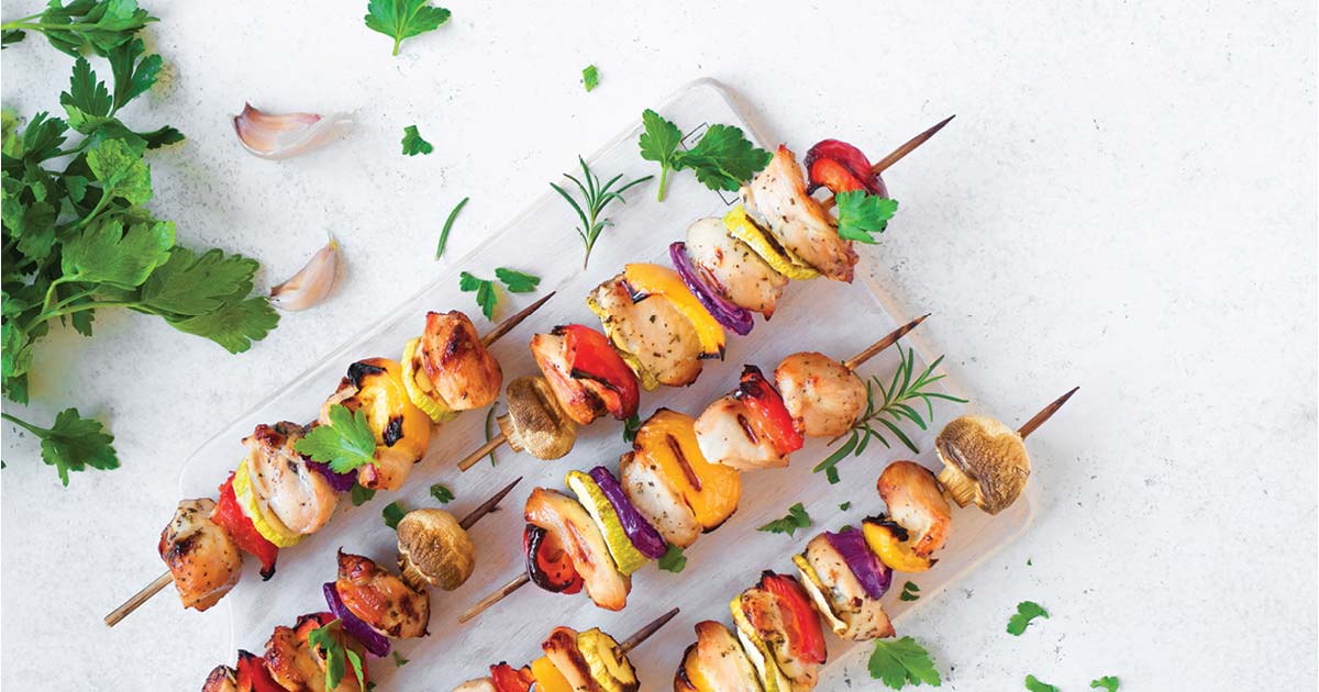Five colorful grilled kebabs, containing pieces of chicken, red onions, bell peppers, and mushrooms, are neatly arranged on a wooden cutting board. Garnished with fresh parsley and sprigs of rosemary, fresh herbs and garlic cloves are scattered around the board.