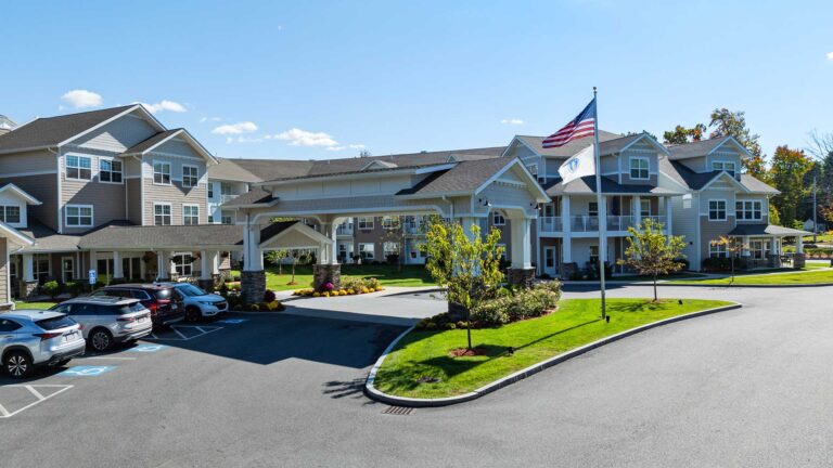 A modern residential building with white and beige siding. It features a covered entrance and well-maintained landscaping. An American flag and another flag are in front. Several cars are parked nearby under a clear blue sky.