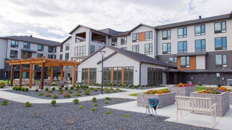 A modern multi-story building with large windows and contrasting gray and white siding. The foreground features a landscaped garden with paths, benches, flower beds, and a wooden pergola under a partly cloudy sky.