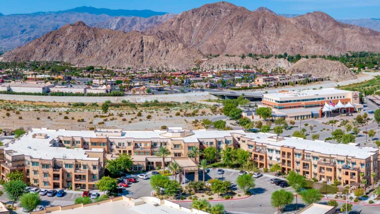 Aerial view of a suburban area featuring light brown residential buildings with white roofs, surrounded by greenery and palm trees. In the background, a large mountain range dominates the landscape under a clear blue sky.