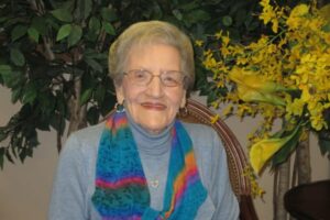 An elderly woman with short gray hair and glasses is smiling while seated. She is wearing a blue turtleneck and a colorful scarf. Behind her, there is a tree and a bouquet of yellow flowers.