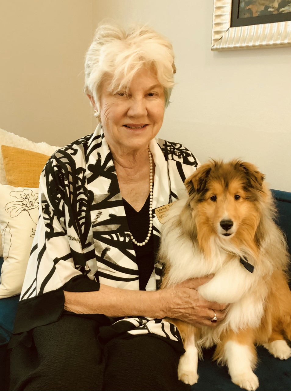 An elderly woman with short white hair sits on a couch, smiling at the camera. She is wearing a black and white patterned jacket and a pearl necklace. She has her arm around a fluffy brown and white dog that is also looking at the camera.