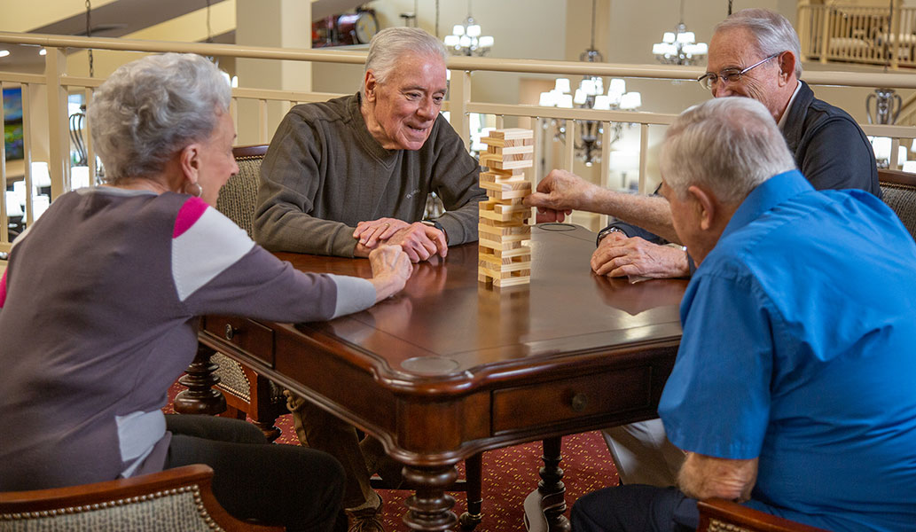 Four elderly adults are sitting around a wooden table playing a game of Jenga. They appear to be in a spacious and well-lit room, engaged and smiling as they take turns carefully removing blocks from the tower. The atmosphere is friendly and relaxed.