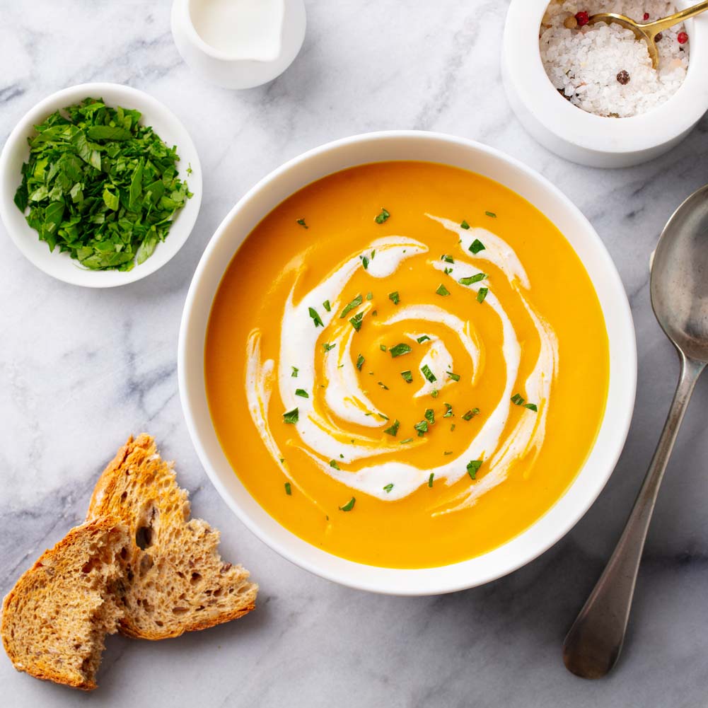 A bowl of creamy pumpkin soup garnished with a swirl of cream and chopped parsley. The soup is placed on a marble surface accompanied by a spoon, a slice of crusty bread, a small bowl of chopped herbs, and a bowl of salt with a spoon.