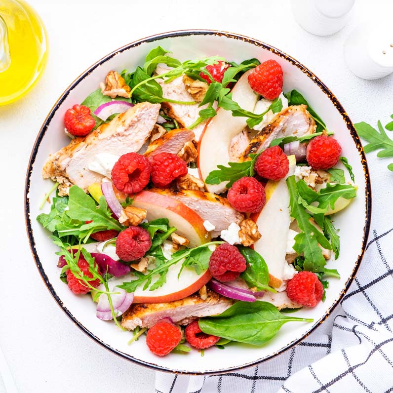 A colorful salad featuring grilled chicken slices, raspberries, apple slices, red onion, walnuts, and mixed greens, served in a white bowl with a brown rim. A striped napkin, a bottle of olive oil, and salt and pepper shakers are nearby.