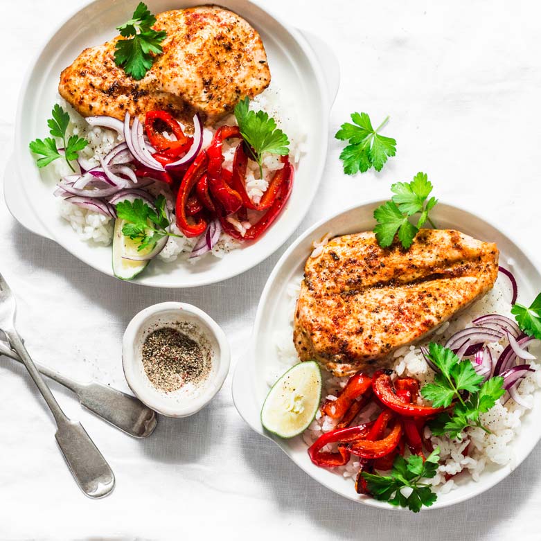 Two white bowls contain grilled chicken breast, sliced red onions, sliced red bell peppers, and cilantro over a bed of white rice. A lime wedge is placed in each bowl. Forks and a small bowl of pepper are beside the dishes on a white tablecloth.