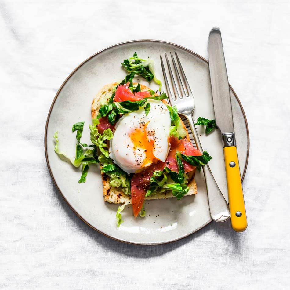 A white plate holds a slice of toast topped with mashed avocado, greens, slices of tomato, and a poached egg with a runny yolk. A fork and knife with yellow handles are placed beside the toast on the plate. The plate sits on a white tablecloth.