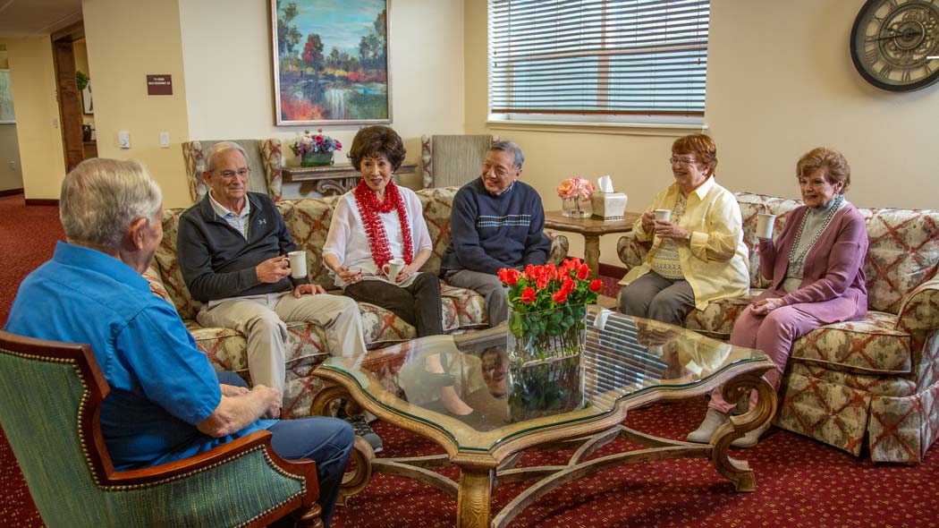 A group of six elderly people, three men and three women, are seated in a cozy living room with patterned couches and a glass coffee table. They are engaged in conversation and enjoying drinks, with a vase of red flowers adding a touch of color to the scene.