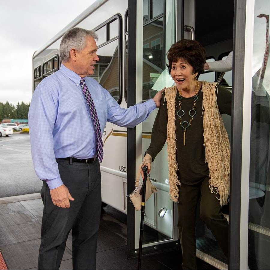 A man in a light blue shirt and striped tie helps an elderly woman with short dark hair, dressed in olive green clothing and holding a cane, as she steps off a bus. The woman appears happy and engaged in conversation.