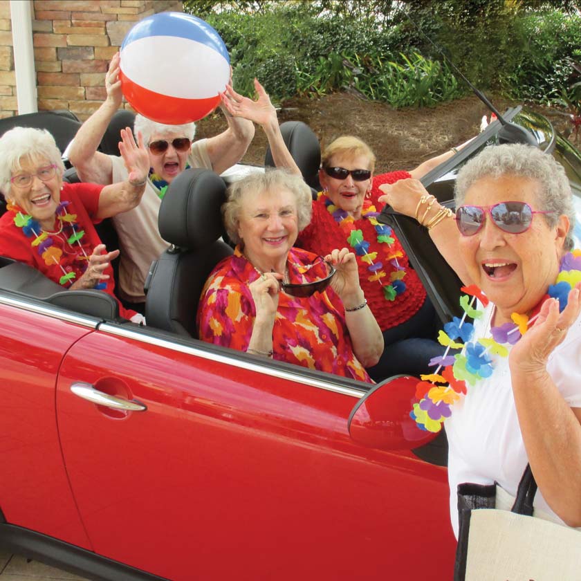 A group of smiling elderly women are seated in a red convertible. They are wearing colorful leis and holding a beach ball, appearing to be enjoying a festive outing. One woman stands outside the car, waving enthusiastically. Greenery and a stone wall are in the background.