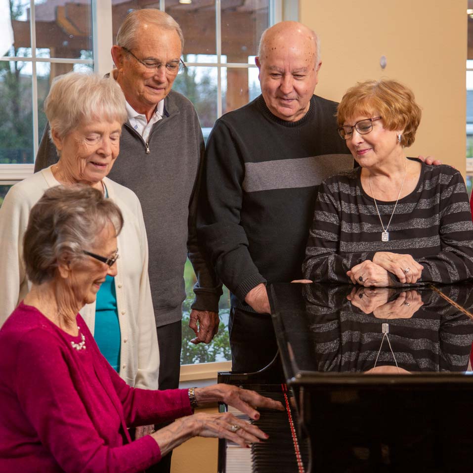 Five elderly people gathered around a grand piano. Four are standing, smiling, and engaged with the woman seated at the piano, who is playing. The group appears cheerful and the setting seems to be a home or community center with large windows in the background.