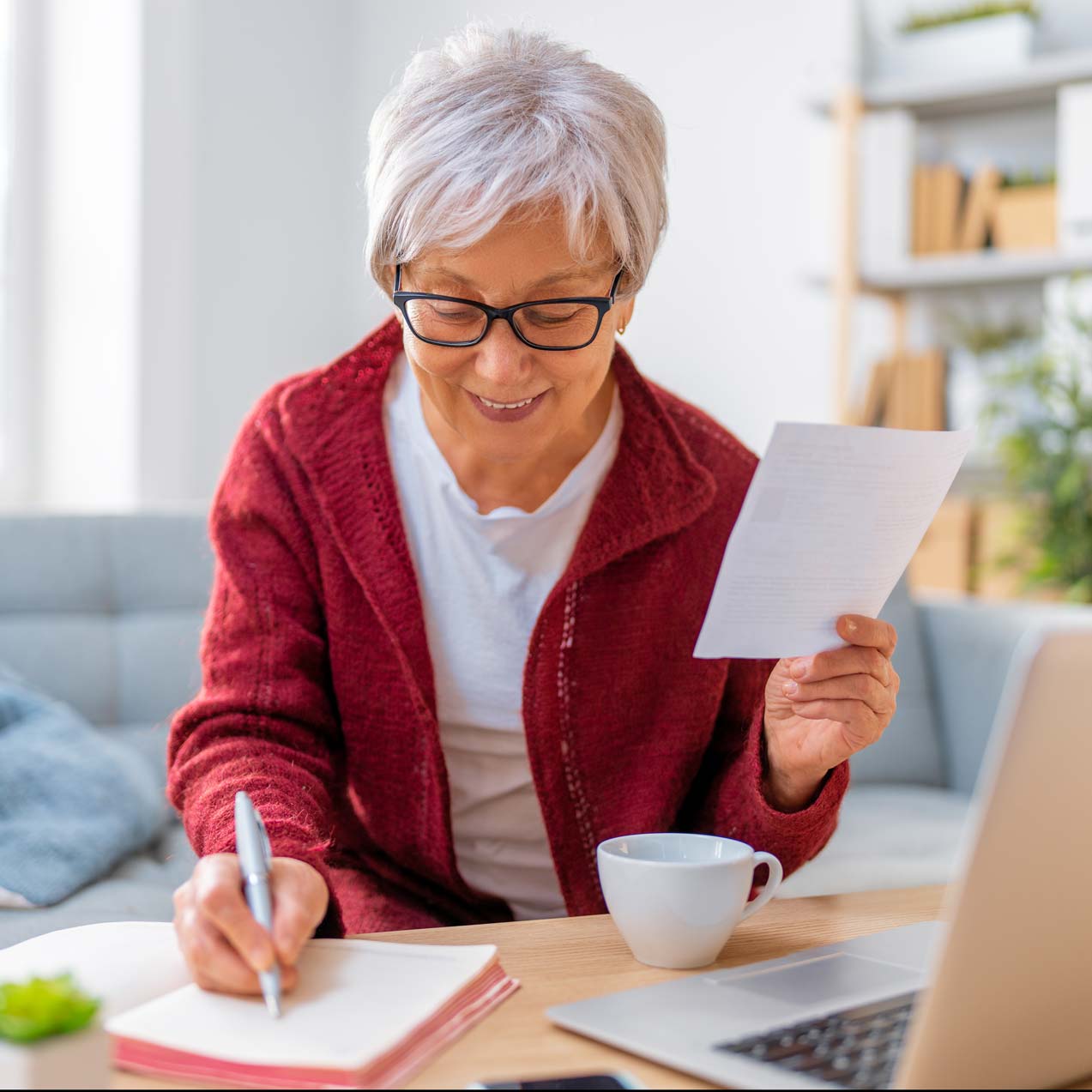 An elderly woman with short grey hair and glasses sits at a table, holding a piece of paper in one hand and writing in a notebook with the other. She is dressed in a red sweater and smiles as she works. A white coffee cup and a laptop are on the table.