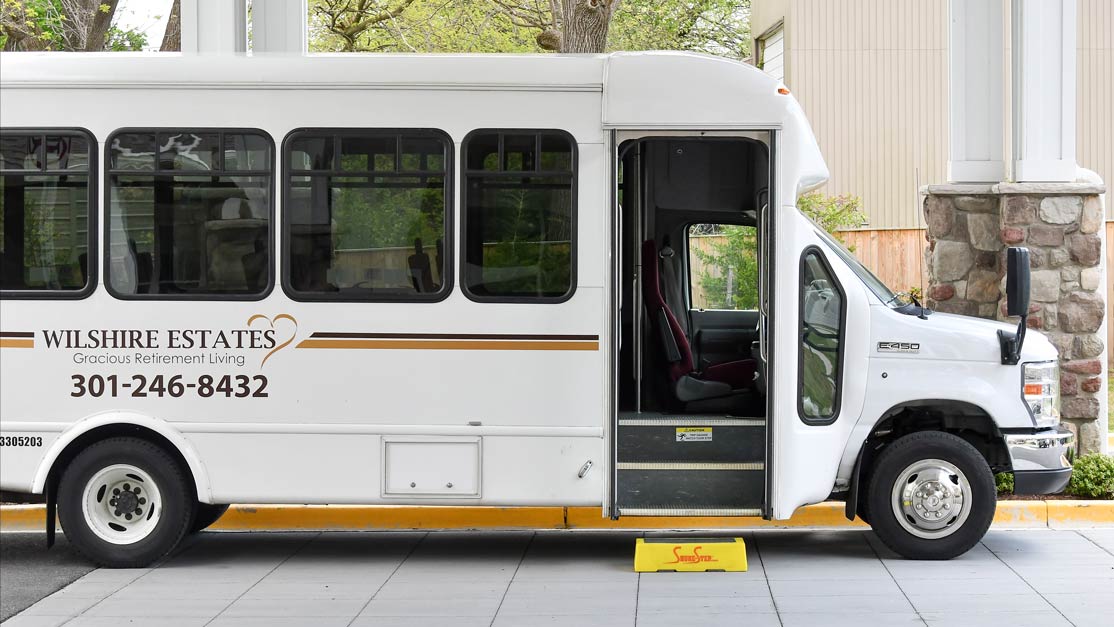 A white shuttle bus with "Wilshire Estates: Gracious Retirement Living" written on its side is parked. The bus door is open, and a yellow step stool is placed on the ground to assist with entry. The backdrop includes trees and part of a building.