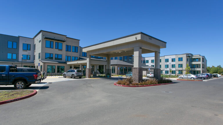 Modern three-story building with a covered driveway entrance, cars parked in front, and clear blue sky in the background. The scene appears to be a residential or medical facility.