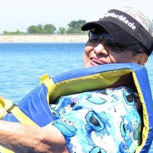 A person is smiling while floating on water wearing a blue life jacket and a cap with sunglasses. They are dressed in a blue shirt with a cartoon character print. The background shows a calm body of water and some greenery in the distance.