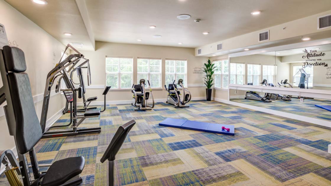 A bright, spacious gym with large windows, featuring exercise equipment like stationary bikes and weight machines. A blue exercise mat with small red dumbbells lies on the floor. Wall mirrors and motivational quotes adorn the space.