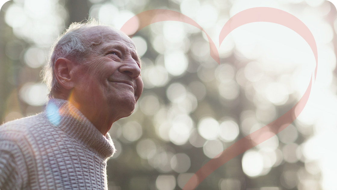 Elderly man in a sweater smiling and looking up while standing outdoors. Soft-focus background with sunlight filtering through trees. A faint, pink heart shape overlays the image on the right side.